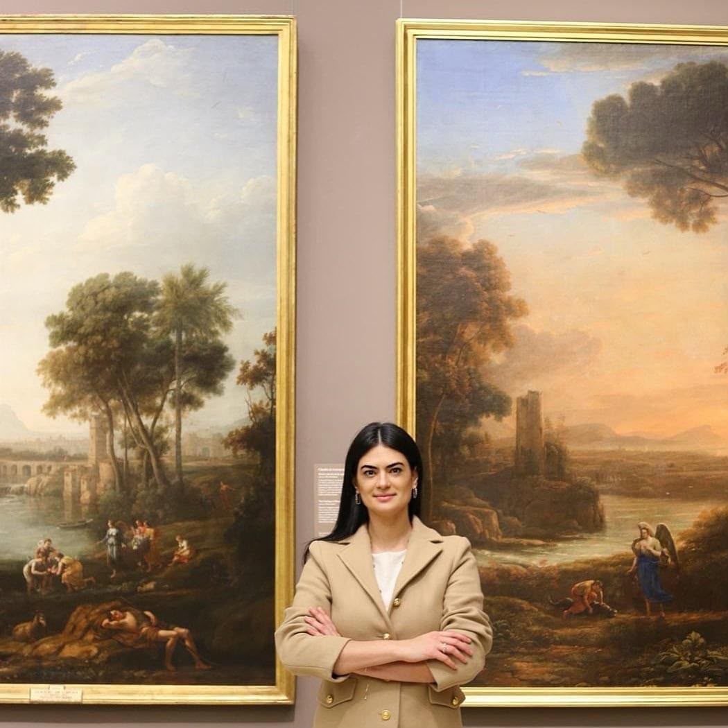 Jhess Alves em um museu diante de duas grandes pinturas de paisagem clássica